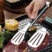 Close-up of stainless steel tongs holding a piece of meat with corn and vegetables in the background.