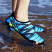 A person wearing blue water shoes with black accents, standing in shallow water at the beach.