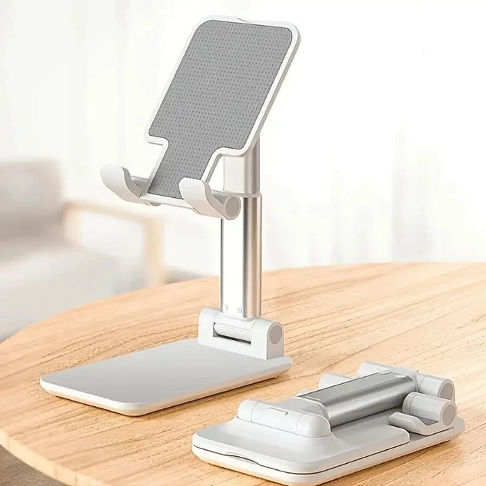 A collapsible phone stand with a sleek, minimalist design. It is primarily white and silver, with a grey, textured grip pad on the phone holder. The stand is shown both in an upright position and collapsed flat on a wooden surface.