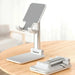 A collapsible phone stand with a sleek, minimalist design. It is primarily white and silver, with a grey, textured grip pad on the phone holder. The stand is shown both in an upright position and collapsed flat on a wooden surface.