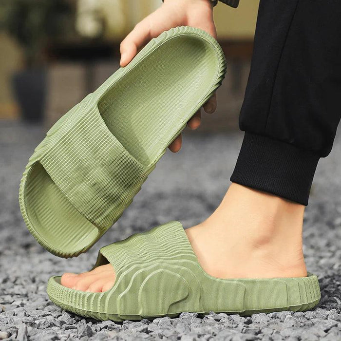 A close-up of a person wearing green slide sandals, holding one sandal in hand with the other foot in the other sandal on a gravel surface.