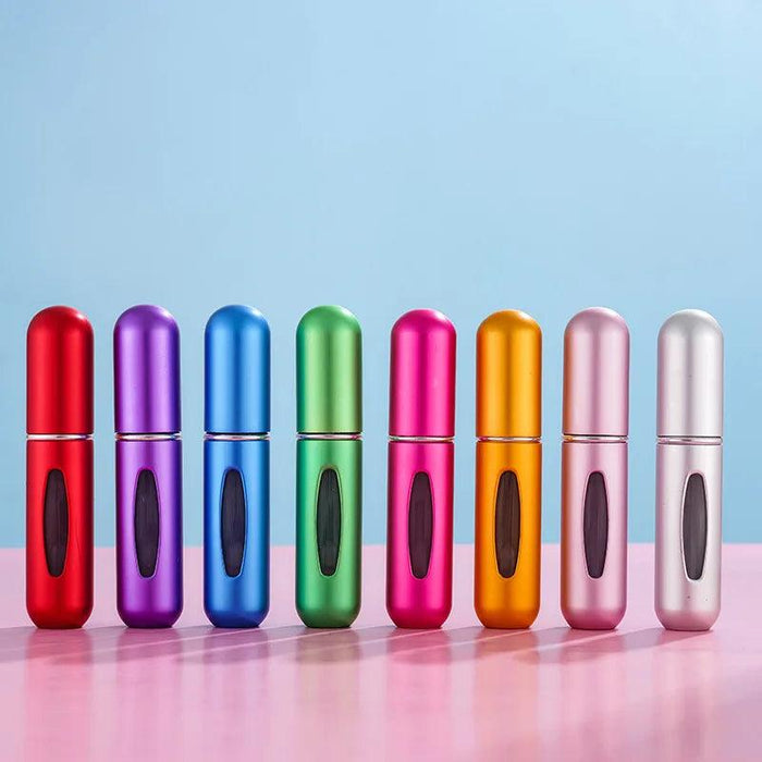 A row of eight portable perfume spray bottles in various colors (red, purple, blue, green, pink, gold, light pink, and silver) on a pink surface with a light blue and light pink background.