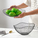 A person holds a black metal wire fruit basket containing several green limes, while a similar empty basket rests on the table below.