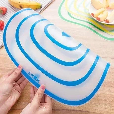 a transparent cutting mat with blue concentric circles. The mat is slightly bent, being held by two hands over a wooden table, with fruits like bananas and strawberries in the background.