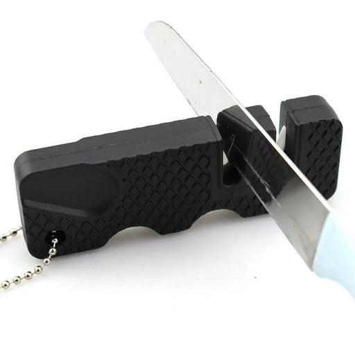 A close-up of a knife being sharpened using a black plastic knife sharpener. The knife is being pulled through one of the slots in the sharpener. The sharpener has a textured surface for better grip and a small chain for attachment, indicating it is designed to be portable and easy to use.