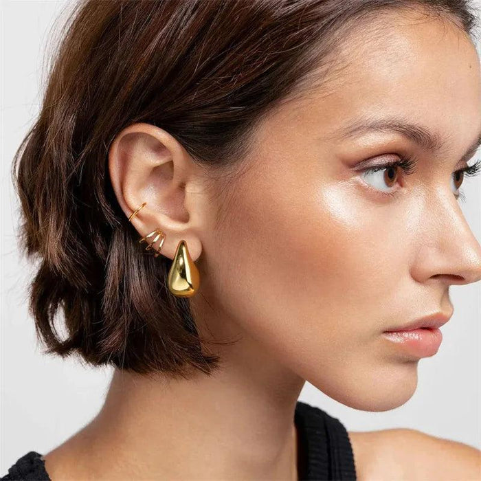 a woman with short brown hair wearing large, teardrop-shaped gold earrings. The earrings are smooth and reflective, giving her a bold and stylish look. She also wears a few smaller, minimalist earrings on the same ear.