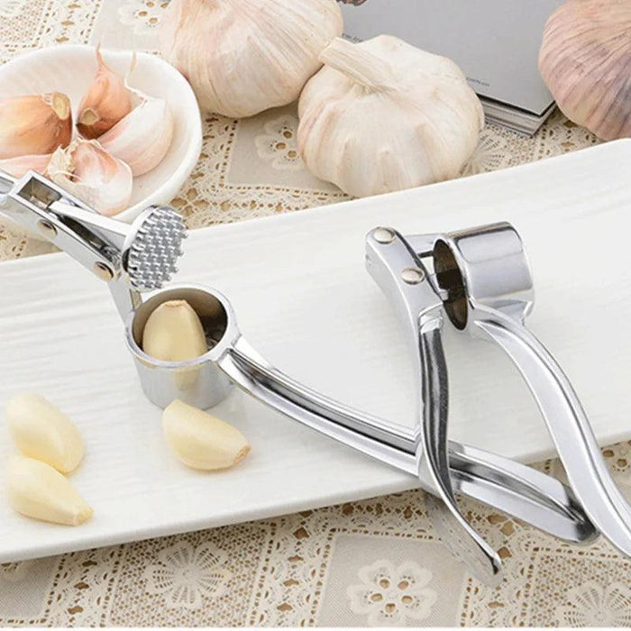 A metal garlic press with a garlic clove inside, surrounded by garlic bulbs and cloves.