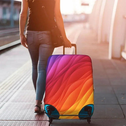 A person walking at a train station with a suitcase covered by a vibrant luggage cover in gradient colors of red, pink, orange, and yellow, resembling a sunset.