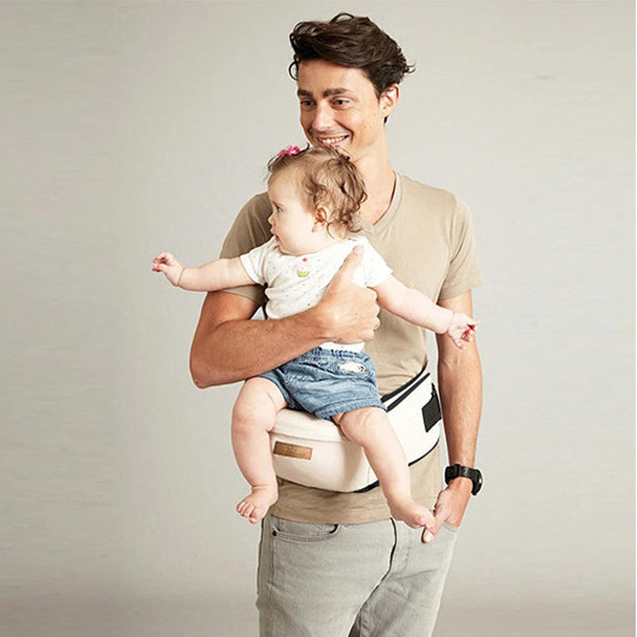 A father smiling while carrying a baby using a cream baby waist carrier, showcasing hands-free support and comfort.