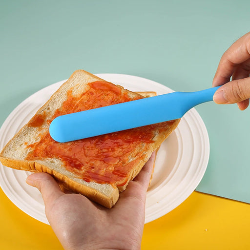 a hand using a blue spatula to spread red jam on a slice of bread over a plate, with a colorful background enhancing the scene.