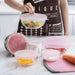 a person placing a silicone stretch lid over a bowl of salad. Several other containers, also covered with silicone lids, are arranged on a kitchen countertop, highlighting the practical use of these lids in everyday food storage.