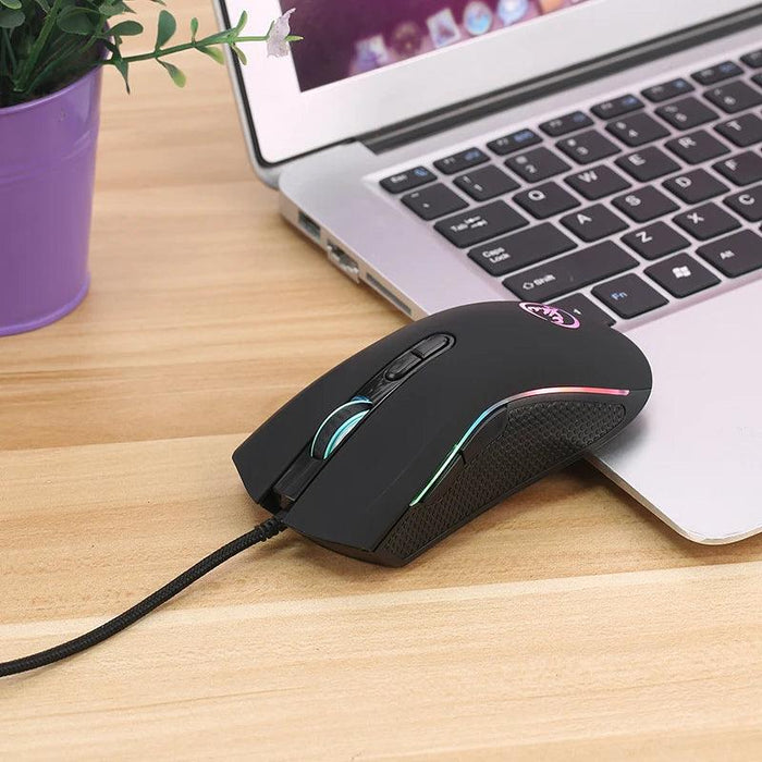 Black gaming mouse from a slightly angled top view. The mouse is placed on a wooden desk next to a laptop. The RGB lighting on the mouse wheel and sides is visible, and the mouse has a smooth, matte finish with a textured grip on the sides.