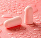 Close-up of two pink earplugs with water droplets on them. Displayed on pink background.