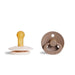 A beige pacifier with a yellow nipple and a brown, round pacifier with a ring handle.