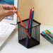 Hand placing an orange pencil into a black mesh pen holder next to a laptop, with additional pens on the desk.