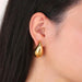 a woman wearing large, teardrop-shaped gold earrings. The earrings have a smooth, polished surface, adding a touch of luxury to her appearance. Her hair is straight and dark, providing a contrast to the shiny gold.