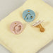 Two round pacifiers with ring handles, one pink and one blue, and a single beige pacifier with a bulbous nipple, are placed on a soft, pale yellow fabric.