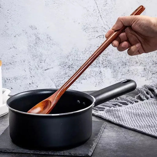 A person stirring a black saucepan with a dark wooden spoon, placed on a dark countertop with a grey cloth in the background.