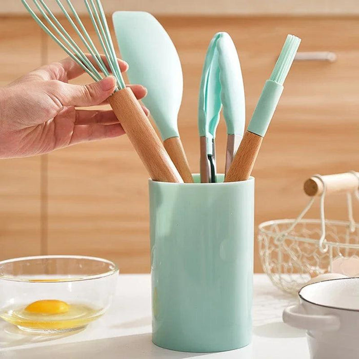 A turquoise silicone kitchen utensil set in a matching holder, on a white surface next to a bowl with an egg and a white basket behind, with a hand taking one utensil from the utensil holder.