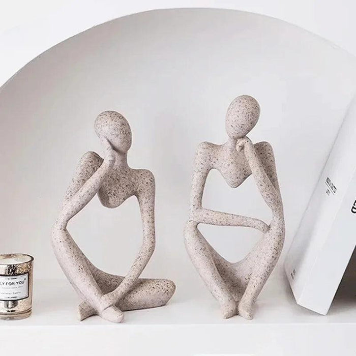 Two abstract sculptures in different reflective poses are displayed on a shelf, accompanied by a candle and a book.