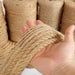 A person holding a bundle of thick jute rope, demonstrating its robust texture and pliability. Several large spools of jute twine are seen in the background, indicating a variety of sizes available for different uses.