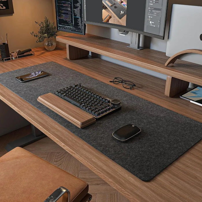 A modern wooden desk setup featuring a large grey desk mat, with a keyboard, mouse, phone, glasses, and dual monitors in the background.