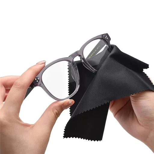 a person cleaning eyeglasses with a black microfiber cloth. One hand holds the glasses, while the other uses the cloth to wipe the lenses, demonstrating the cloth's functionality.