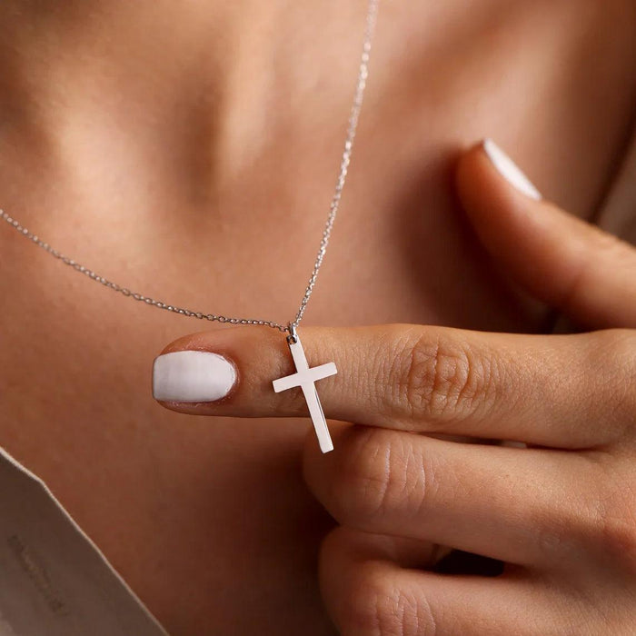 A person holding a silver cross necklace between their fingers, with a close-up view of their neck and part of their light-colored top.