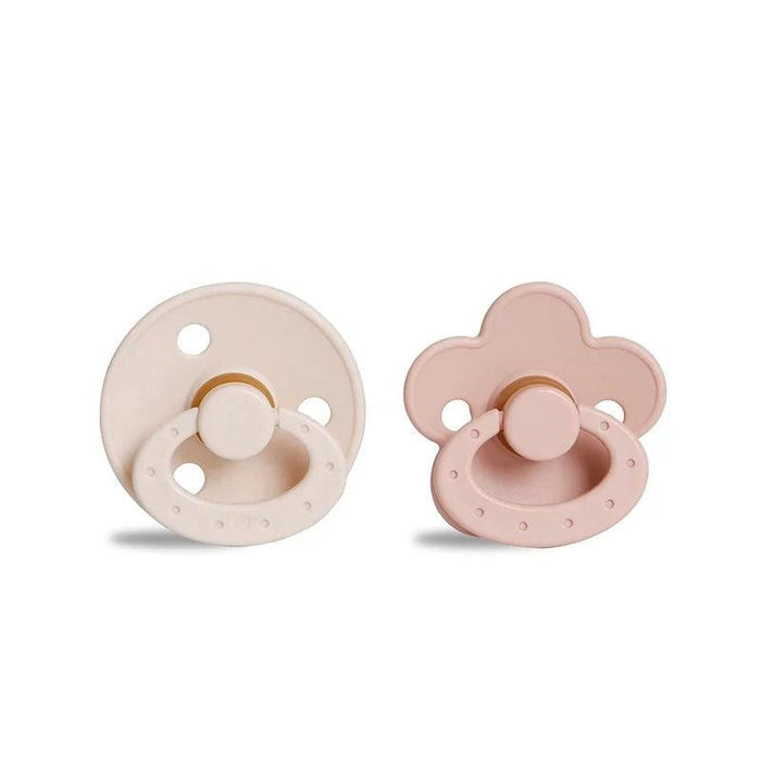 A round, beige pacifier with a ring handle next to a pink, flower-shaped pacifier with a ring handle.