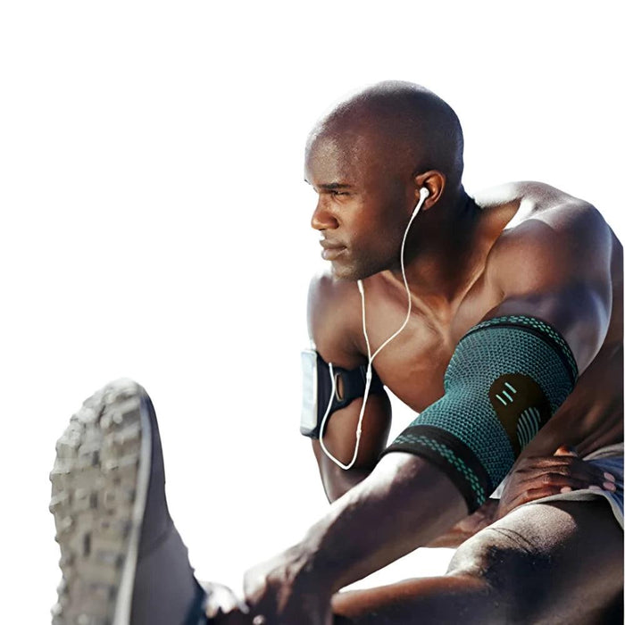 A muscular man with dark skin is stretching his leg while wearing a black elbow support wrap on his arm. He is outside, focused on his exercise, with earphones in his ears and an armband holding a device on his other arm.