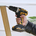 a hand using the yellow and black Yikoda cordless power drill to drive a screw into a wooden piece, showcasing its practical application. The drill runs on a 21V lithium battery, ensuring portability and ease of use.