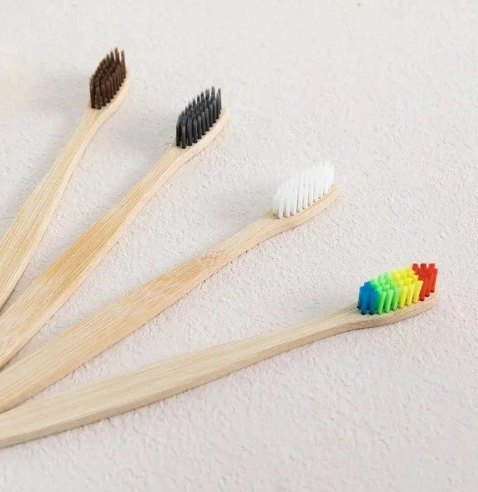 Four bamboo toothbrushes laid out on a light-colored surface. Each toothbrush has a different color bristle: black, white, brown, and multicolored (rainbow).