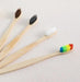 Four bamboo toothbrushes laid out on a light-colored surface. Each toothbrush has a different color bristle: black, white, brown, and multicolored (rainbow).