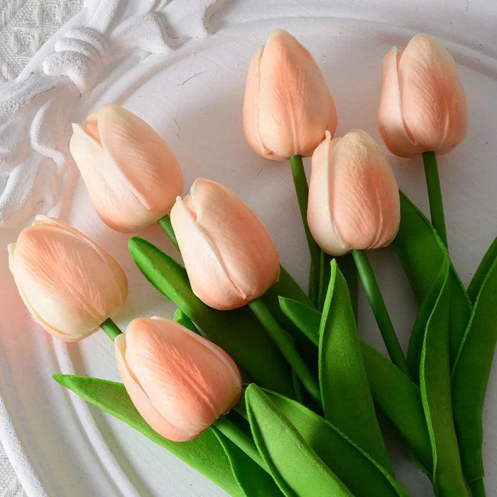 Artificial peach-colored tulips with green leaves on a decorative white plate.