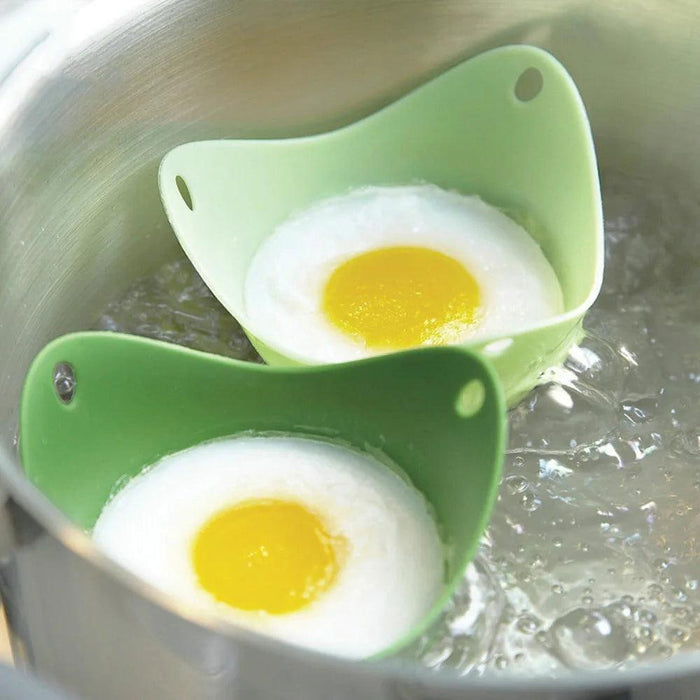 Two green silicone egg poachers, each holding a poached egg, floating in boiling water inside a metal pot.