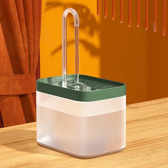 A green pet water fountain with a clear curved spout on a wooden surface, against an orange background. The fountain has a transparent water container.