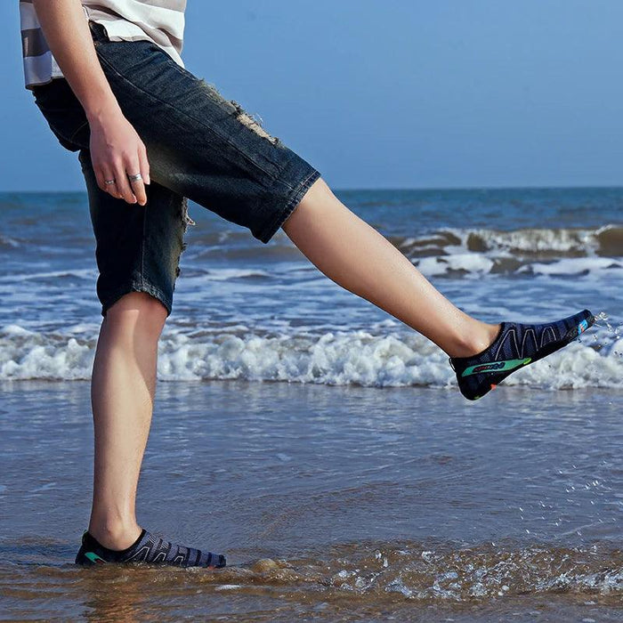 A person wearing black and blue water shoes, standing at the edge of the sea, with the waves in the background.