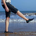 A person wearing black and blue water shoes, standing at the edge of the sea, with the waves in the background.