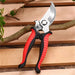 A pair of pruning shears with red and black ergonomic handles, positioned on a wooden surface with a branch in the background.