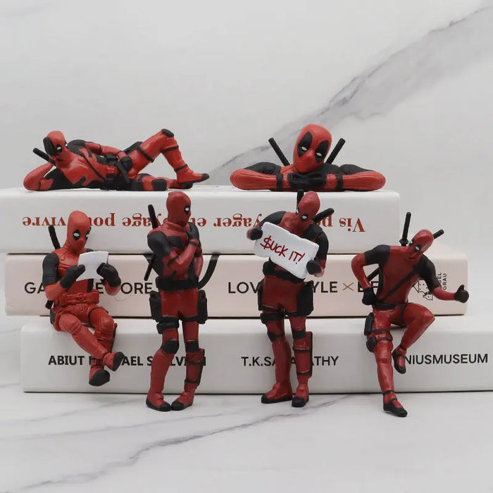 six small Deadpool figurines posed in various positions on top of a stack of books. Each figurine has a different stance, including sitting, leaning, and holding a sign that says "Suck it!"