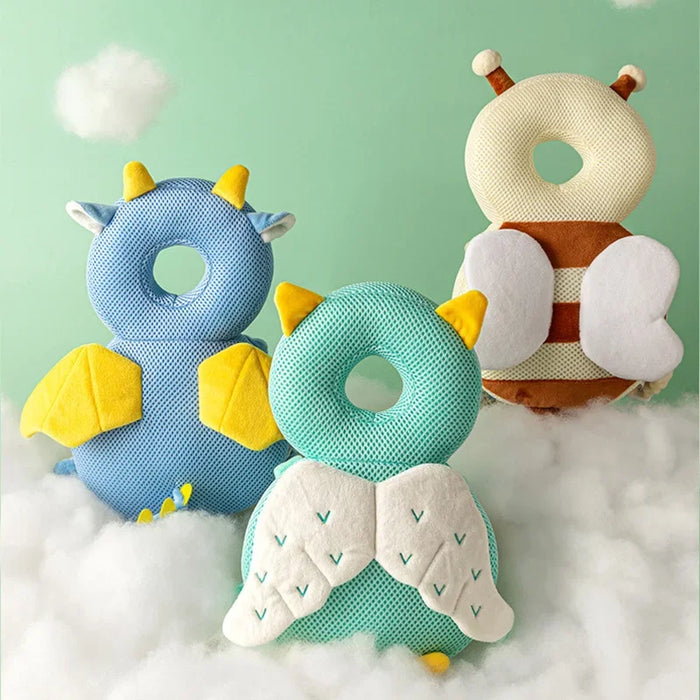 Three baby head protectors displayed on a fluffy white surface: a blue dragon with yellow wings and horns, a green owl with yellow feet and detailed wings, and a bee with brown stripes and white wings.