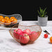 A gold wire fruit basket holding several red apples, placed on a marble table. In the background, a white wire basket filled with oranges and a small potted plant are also visible, adding a touch of greenery to the modern setup.