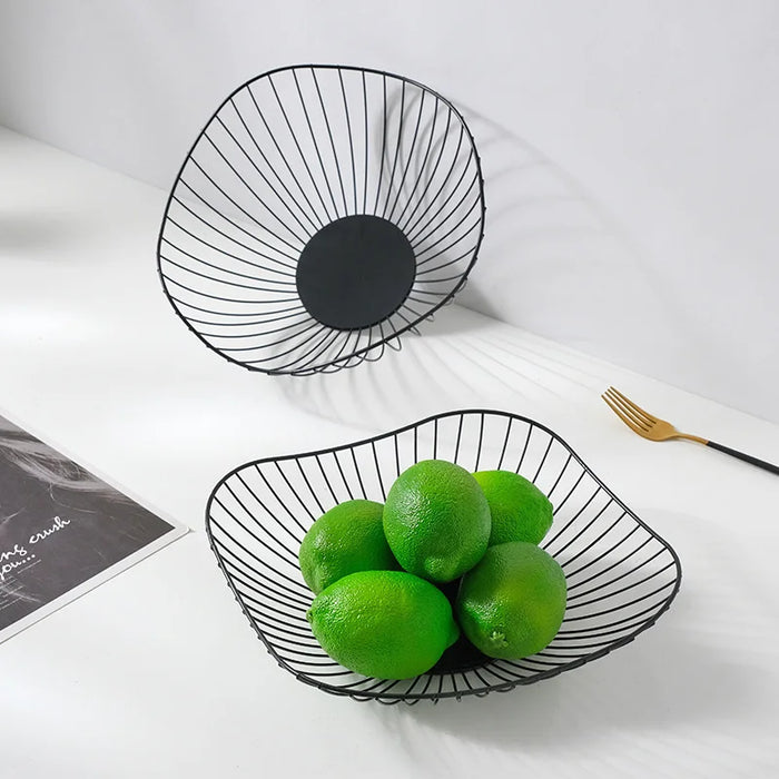 Two black metal wire baskets are placed on a white surface; one is filled with green limes, and the other is empty. A gold spoon and a magazine are also on the table.