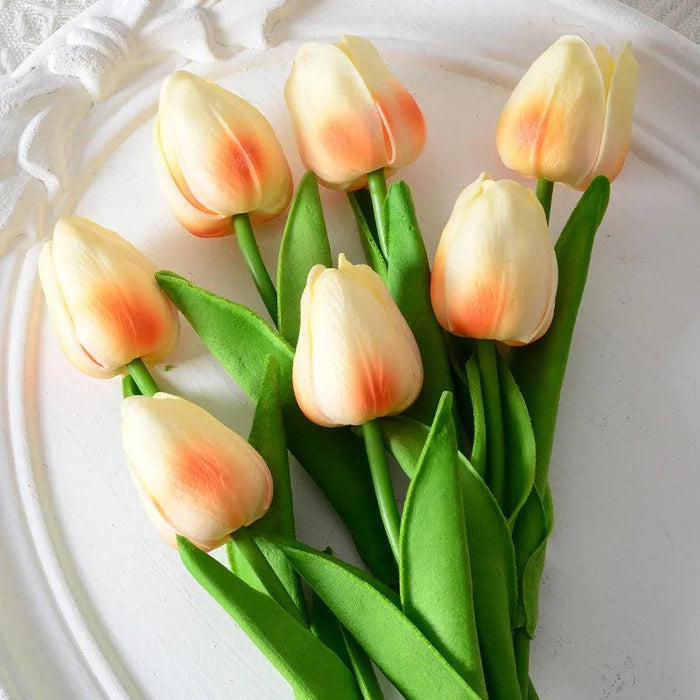 A bunch of artificial tulips with cream and orange petals and green leaves on a white plate.