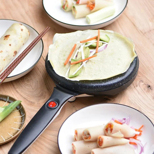 The black electric crepe maker with a finished crepe topped with sliced vegetables, surrounded by plates of prepared crepes and fillings.