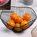 A close-up of a black wire fruit basket filled with bright oranges and a few green apples, sitting on a marble tabletop. This image highlights the basket's open design, perfect for displaying fresh fruits.