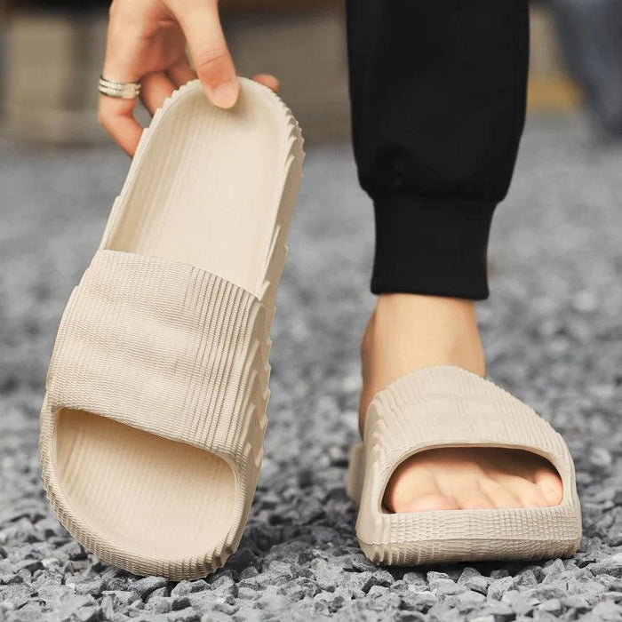 A close-up of a person wearing beige slide sandals, holding one sandal in hand with the other foot in the other sandal on a gravel surface.