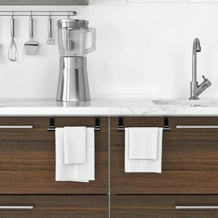 a sleek, stainless steel kitchen counter with a modern, minimalistic blender and hanging kitchen tools in the background. Below the counter, there are two wooden cabinet doors with over-the-door towel racks in black, each holding a white towel. The setup suggests an organized and functional kitchen space.