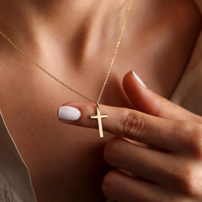 A person holding a gold cross necklace between their fingers, with a close-up view of their neck and part of their light-colored top.