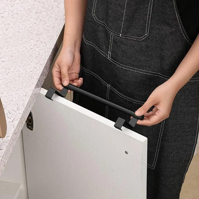 a person installing a black over-the-door towel rack on a white cabinet door. The person is wearing a black apron, indicating a kitchen setting. The image demonstrates the ease of installation of the towel rack, which does not require any tools.